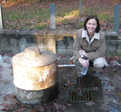 A young woman squats at the side of the spring, filling a plastic water bottle.